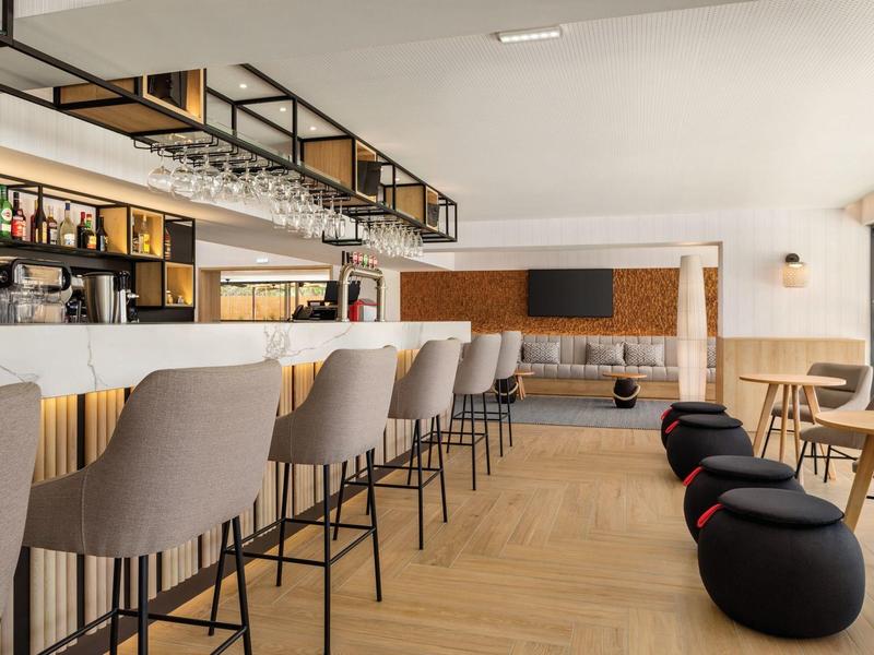 Innenraum einer modernen Bar mit hohen grauen Barhockern, Holztischen und schwarzen Sitzkissen.