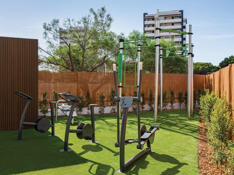 Outdoor-Fitnessgeräte auf grünem Kunstrasen, umgeben von Holzsichtschutz und Bäumen.