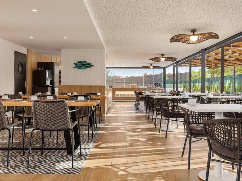 Geräumiger Restaurantinnenraum mit Holzboden, modernen Korbstühlen und großen Glasfenstern.