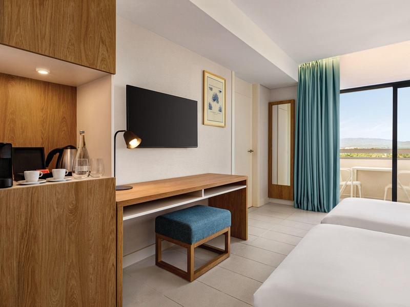 Modernes Hotelzimmer mit Holzmöbeln, TV, Schreibtisch, blauen Vorhängen und Balkon mit Weitblick.