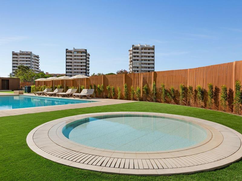 Poolbereich mit rundem Kinderbecken, Liegestühlen, grünem Rasen und Hochhäusern im Hintergrund.