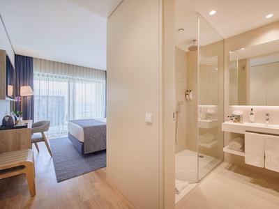 Helles Hotelzimmer mit Holzfußboden, Doppelbett, Schreibtisch und angrenzendem Bad mit Glasdusche.