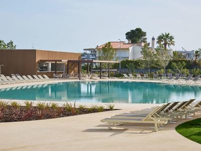 Großer, klarer Pool mit umliegenden Liegestühlen, grüner Rasen und Häuser im Hintergrund.