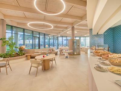 Moderner, heller Speisesaal mit großen Fenstern, runden Tischen, Stühlen und Buffetbereich.