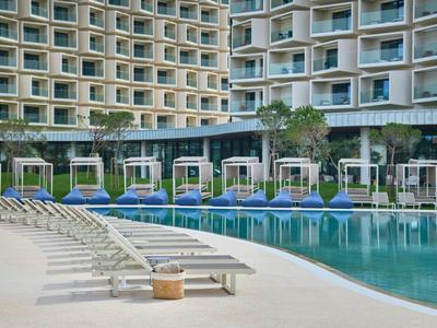 Großer Hotelpool mit weißen Liegen und blauen Schirmen vor mehrstöckigem Gebäude.