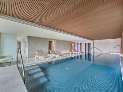 Innenraum eines Swimmingpools mit Holzdach, Liegestühlen und großer Fensterfront.