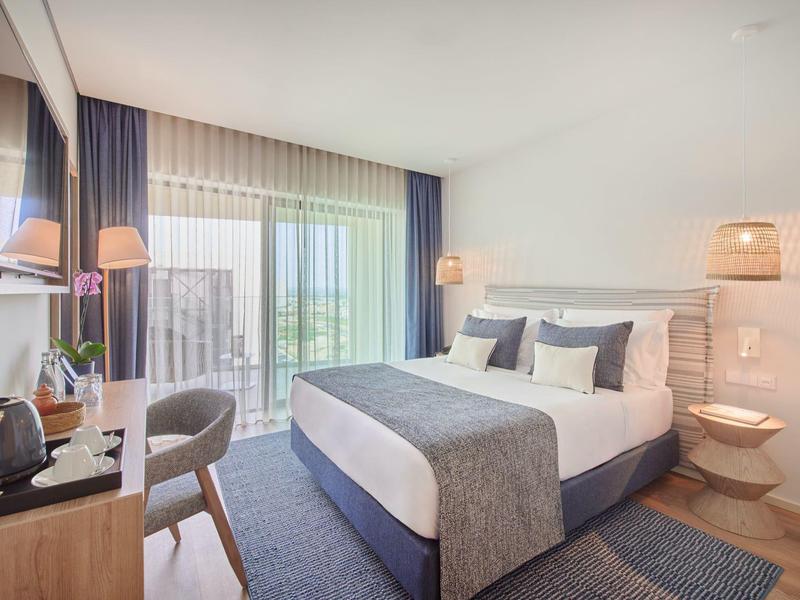 Helles Hotelzimmer mit großem Bett, grauem Teppich, Schreibtisch, Stuhl und Balkon mit grüner Aussicht.