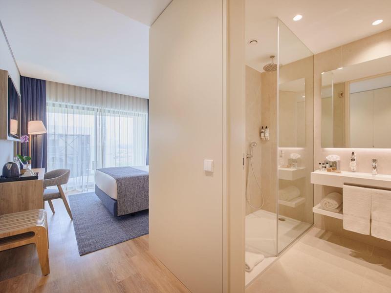 Helles Hotelzimmer mit Holzfußboden, Doppelbett, Schreibtisch und angrenzendem Bad mit Glasdusche.