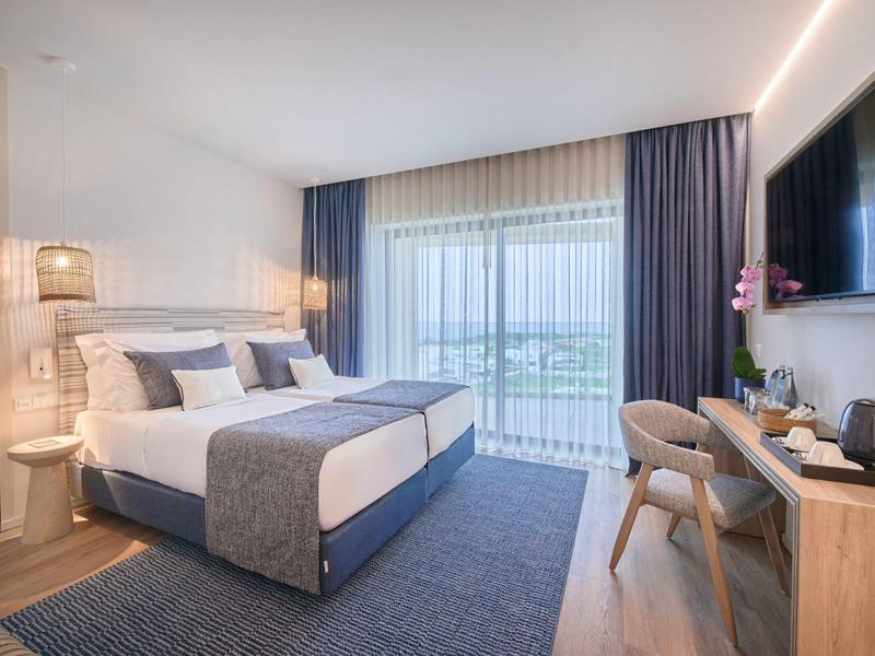 Helles Schlafzimmer mit Doppelbett, blauen Vorhängen, großem Fenster und modernem Holzmobiliar.