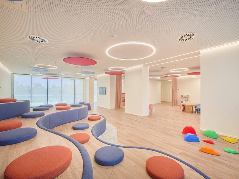 Heller, moderner Raum mit Holzfußboden, bunten Sitzkissen und runden Deckenleuchten.