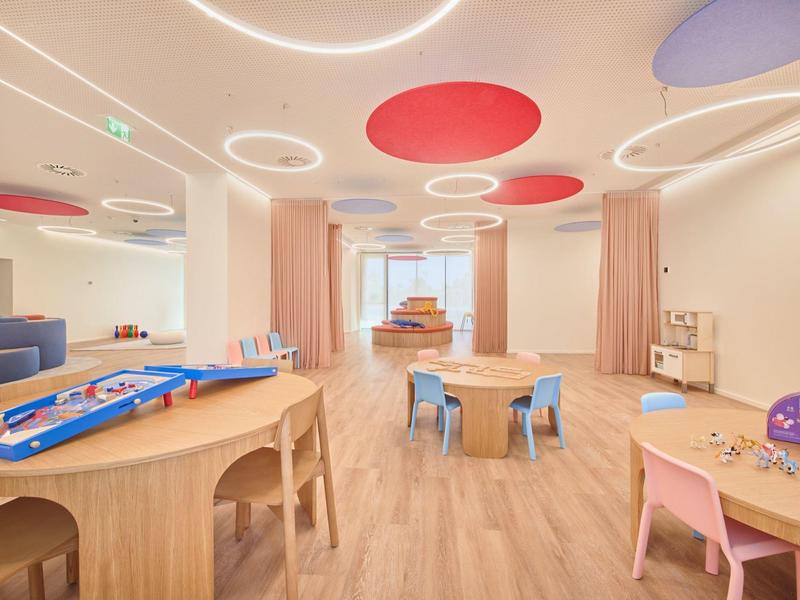 Heller Raum mit Holzmöbeln, bunten Deckenpanelen und Spieltischen in Kindergartenumgebung.