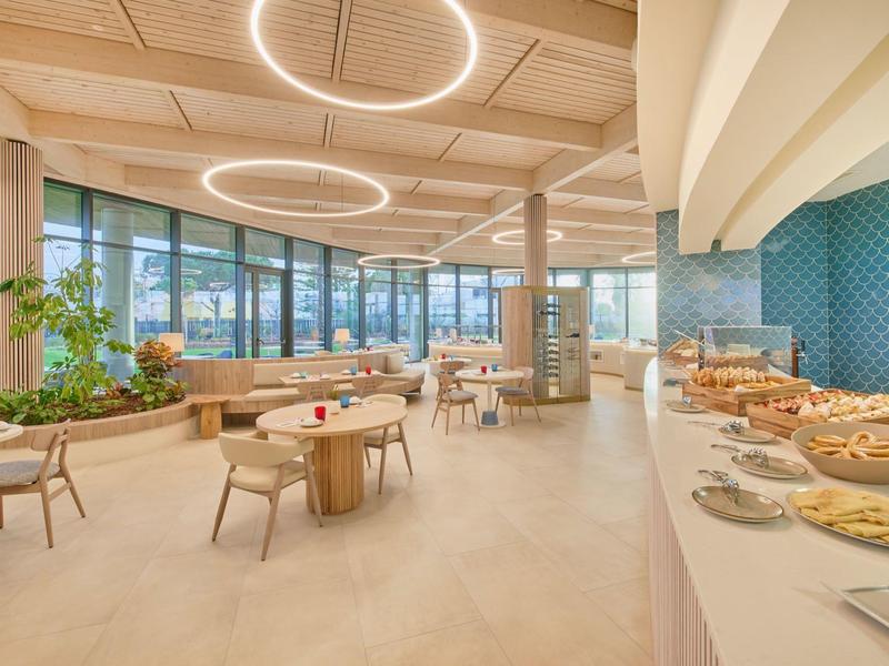 Moderner, heller Speisesaal mit großen Fenstern, runden Tischen, Stühlen und Buffetbereich.