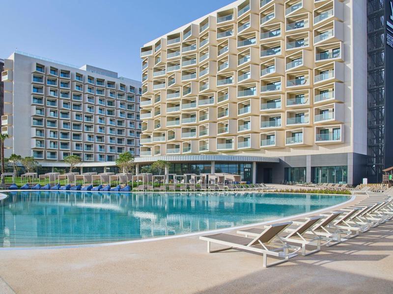 Großes Hotel mit zwei Gebäuden, blauer Pool, Liegestühle und sonniger Himmel.
