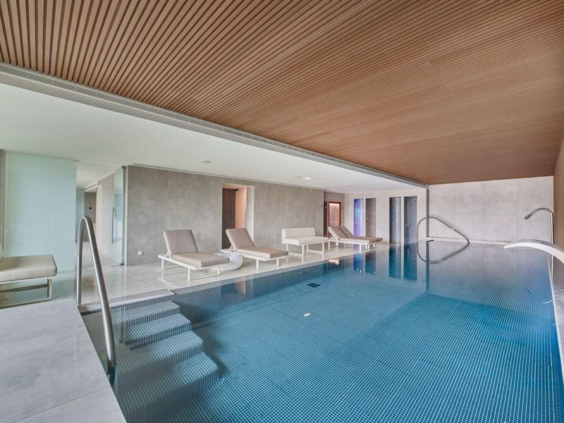 Innenraum eines Swimmingpools mit Holzdach, Liegestühlen und großer Fensterfront.