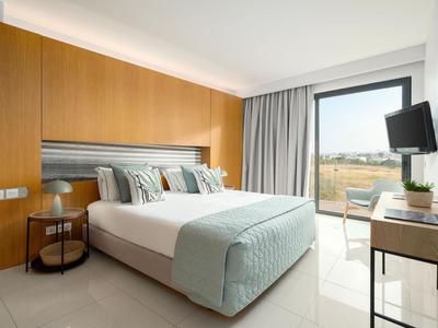 Heller Hotelzimmer mit großem Bett, Holzakzenten und Fenster mit Blick auf Außenbereich.