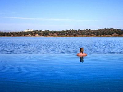 Frau entspannt im Infinity-Pool mit Blick auf See und Baumlinie unter blauem Himmel.