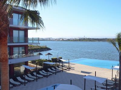 Hotelterrasse mit Liegestühlen, Palmen und Infinity-Pool mit Blick auf einen See bei klarem Himmel.