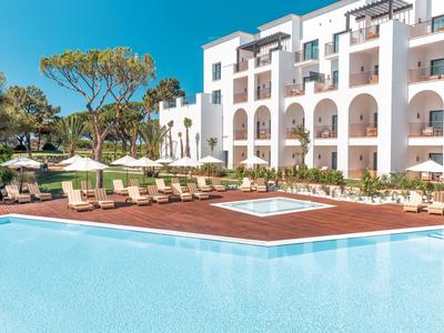 Großer blauer Pool mit Holzdeck und weißen Liegestühlen vor einem weißen Hotelgebäude.