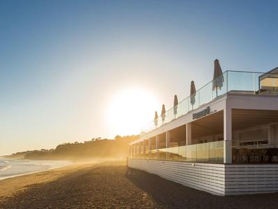 Strand mit Sand, blauem Himmel und sonnigem Morgenlicht, neben einem modernen Café mit Terrasse.