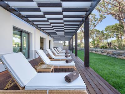 Eine Terrasse mit mehreren weißen Liegestühlen auf Holzlatten unter einem Pergoladach neben einem grünen Rasen.