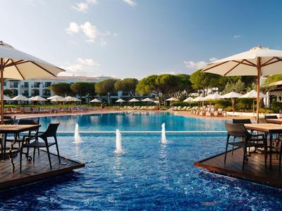Großer blauer Pool mit Springbrunnen, umgeben von Sitzbereichen mit Sonnenschirmen und Lounge-Stühlen.