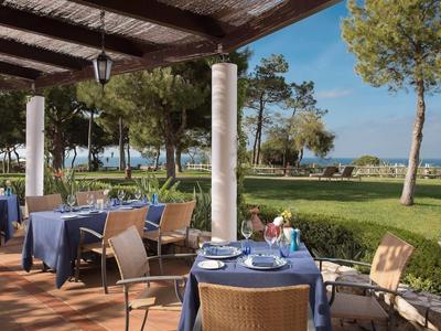 Terrassenbereich mit gedeckten Tischen unter Holzüberdachung, Blick auf Rasen, Bäume und Meer.