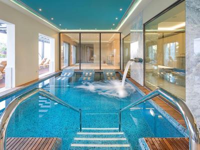 Innenbereich mit blauem Schwimmbecken, Wasserdüsen, Holzterrasse und großen Fenstern.