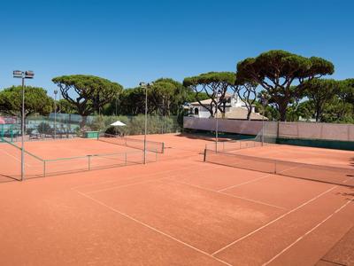 An empty red clay tennis court with green trees in the background under a clear blue sky.
