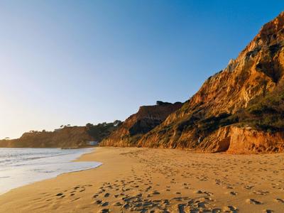 Strand mit goldenem Sand, ruhigem Meer und steilen Felsen unter klarem, blauem Himmel bei Sonnenuntergang.