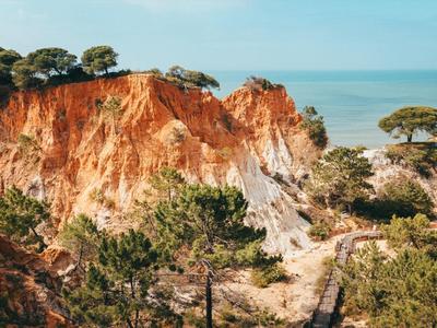 Rote Felsenklippe mit grünen Bäumen vor blauem Himmel und Meer im Hintergrund.