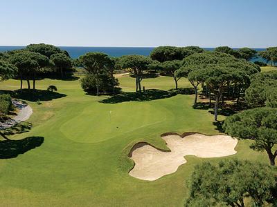 Golf course with sand bunker, surrounded by trees and a view of the sea in the background.
