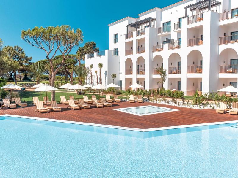 Großer blauer Pool mit Holzdeck und weißen Liegestühlen vor einem weißen Hotelgebäude.