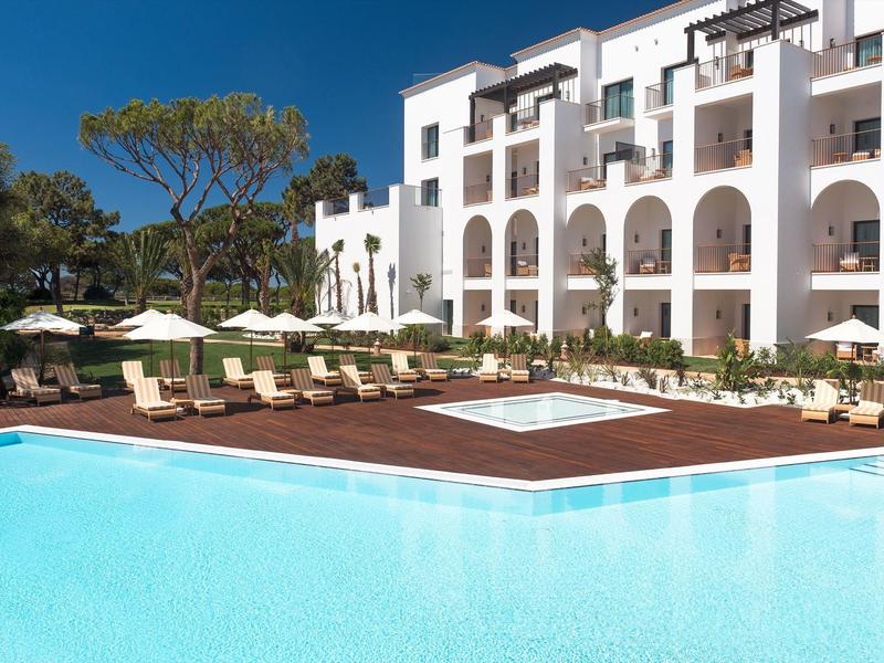 Großer blauer Pool vor weißem Hotel mit Arkaden und Liegestühlen unter Sonnenschirmen.