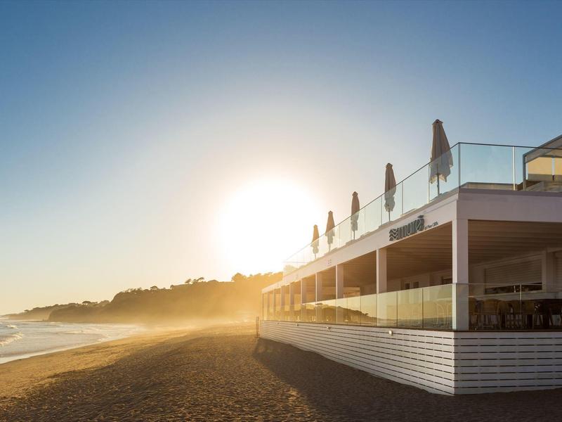 Strand mit Sand, blauem Himmel und sonnigem Morgenlicht, neben einem modernen Café mit Terrasse.