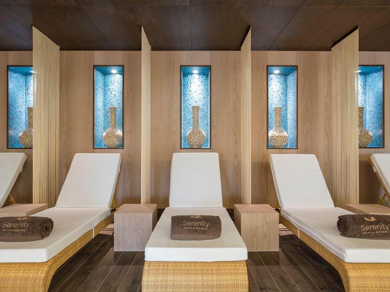 Ruhebereich mit weißen Liegen, Holzwänden und dekorativen Nischen mit blauen Glasflaschen.