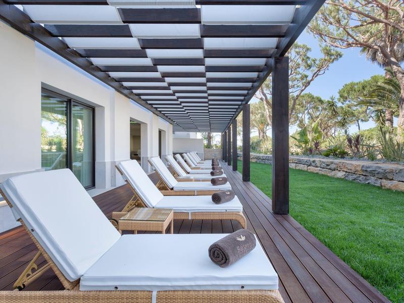 Eine Terrasse mit mehreren weißen Liegestühlen auf Holzlatten unter einem Pergoladach neben einem grünen Rasen.