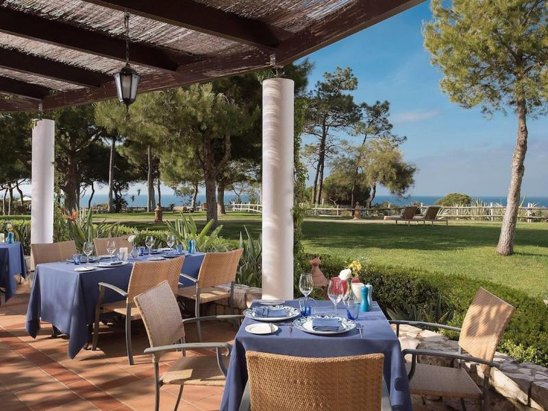 Terrassenbereich mit gedeckten Tischen unter Holzüberdachung, Blick auf Rasen, Bäume und Meer.