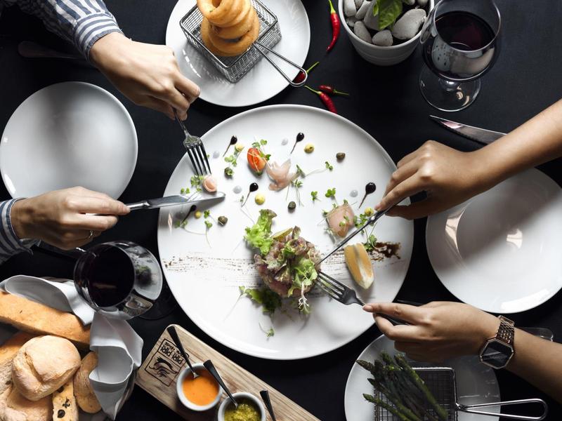 Menschen essen gemeinsames Gericht mit Fleisch, Salat, Brot und Getränken auf dunklem Tisch.