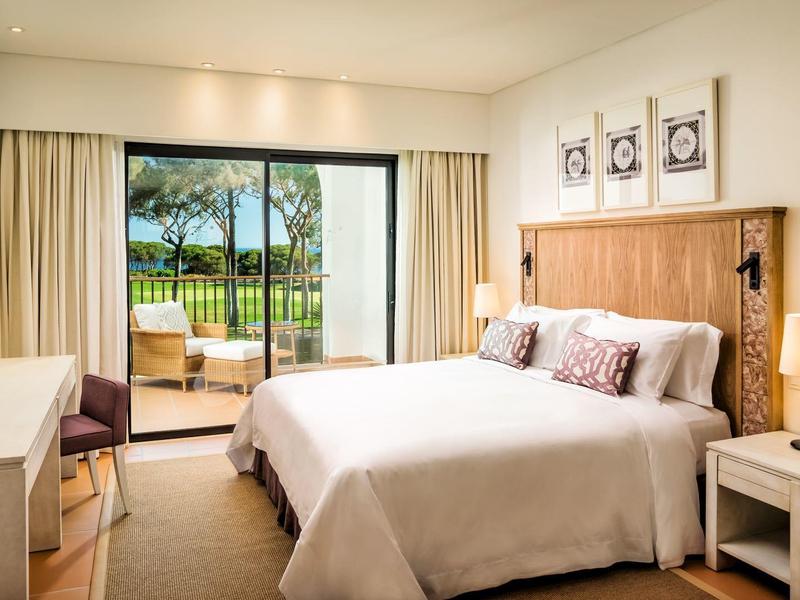 Helles Schlafzimmer mit großem Bett, Holzmöbeln und Blick auf Balkon mit Grünfläche und See.