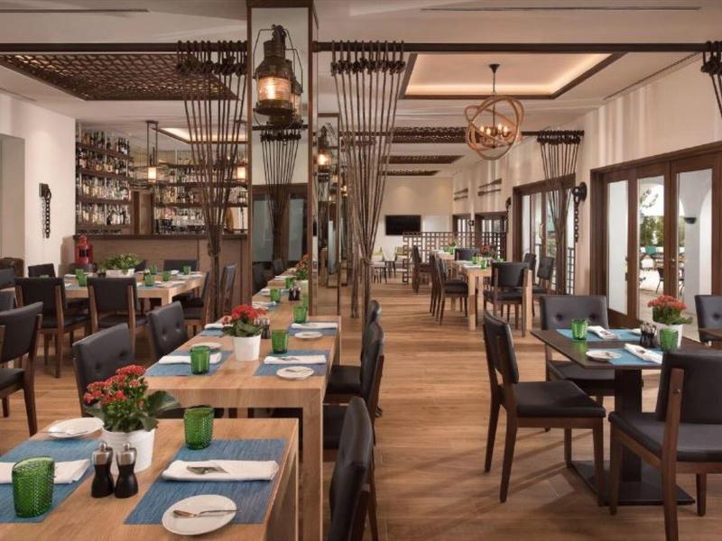 Modernes Restaurant mit Holzböden, grünen Pflanzen, dunklem Mobiliar und großem Fensterfronten.