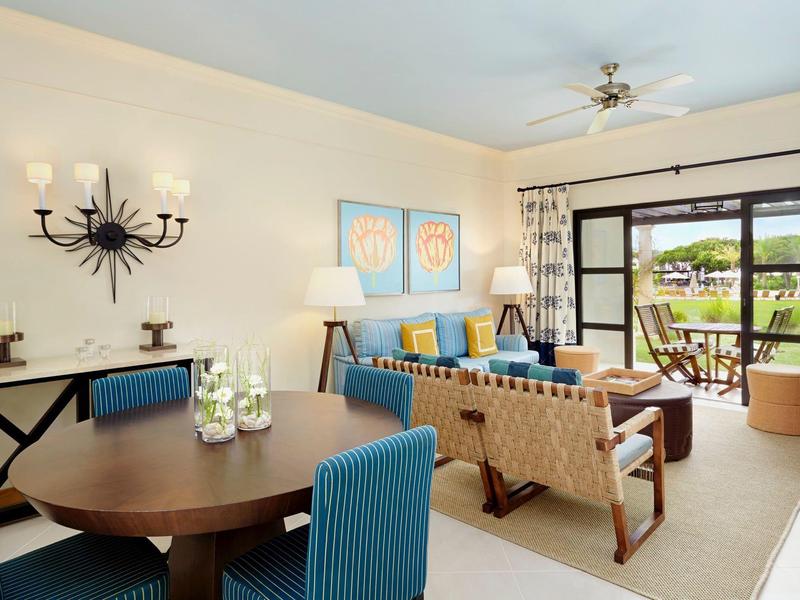 Helles Wohnzimmer mit runden Holztisch, blauen Stühlen, Sofa, großer Fensterfront und Gartenausblick.