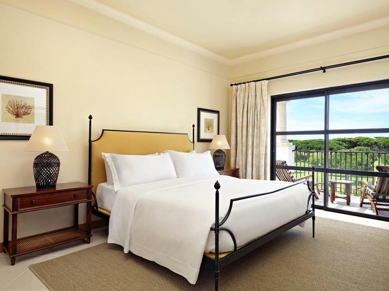 Helles Schlafzimmer mit großem Bett, Holzmöbeln, Kunstbildern und Balkon mit grünem Ausblick.