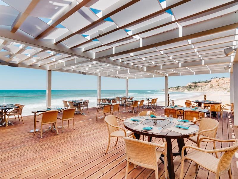 Offenes Strandrestaurant mit Holzterrasse, gedeckten Tischen und Blick auf das blaue Meer.