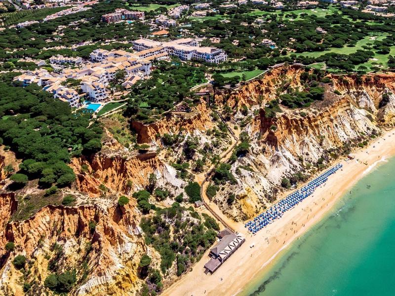 Lange Sandküste mit steilen roten Klippen, grüner Vegetation und blauem Meer unter klarem Himmel.