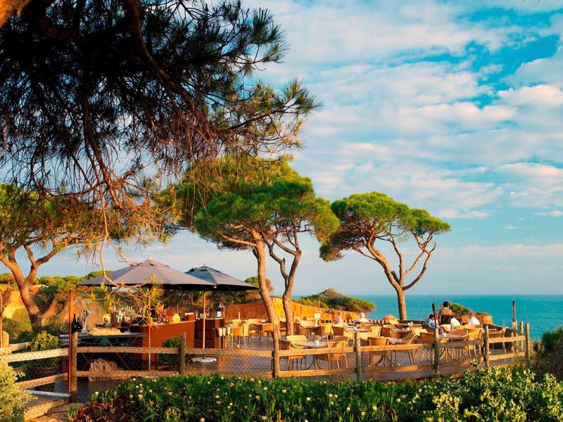 Strandrestaurant mit Tischen, Stühlen und Bäumen vor blauem Himmel und Meer im Hintergrund.