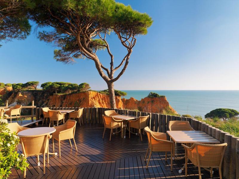 Terrasse mit Stühlen und Tischen mit Meerblick unter blauem Himmel und Bäumen.