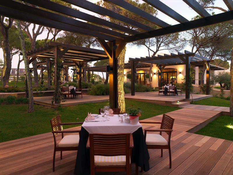 Terrasse mit Holzboden, gedecktem Tisch und vier Stühlen, umgeben von Bäumen und beleuchteten Gebäuden.