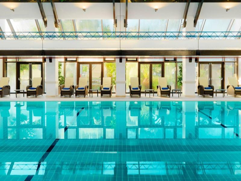 Schwimmbad mit klarem blauem Wasser, hohen Decken und Glasfenstern, Umkleidekabinen im Hintergrund.