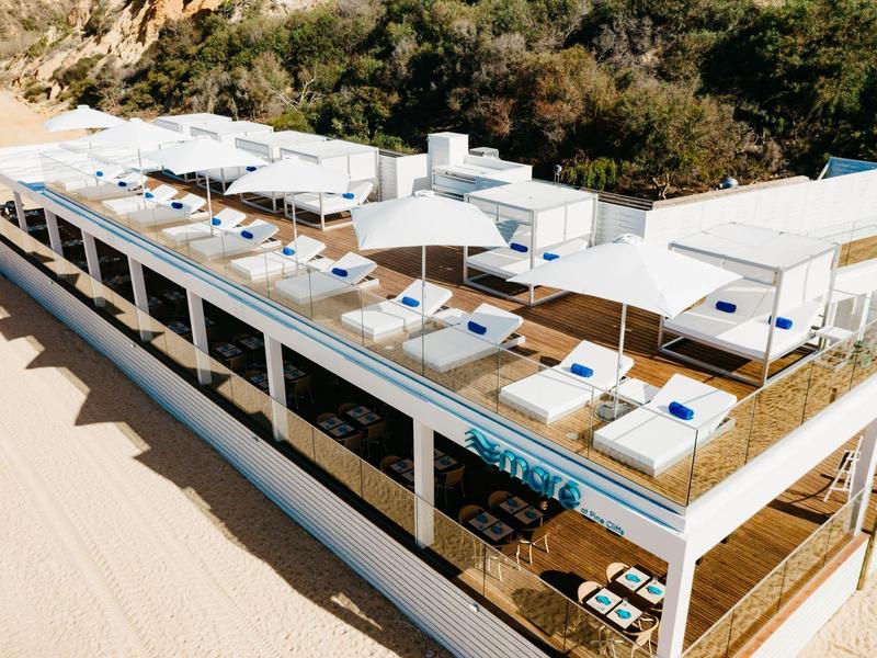 Modernes, rechteckiges Strandrestaurant mit weißem Dach und Sitzbereichen, umgeben von Sand und Dünen.