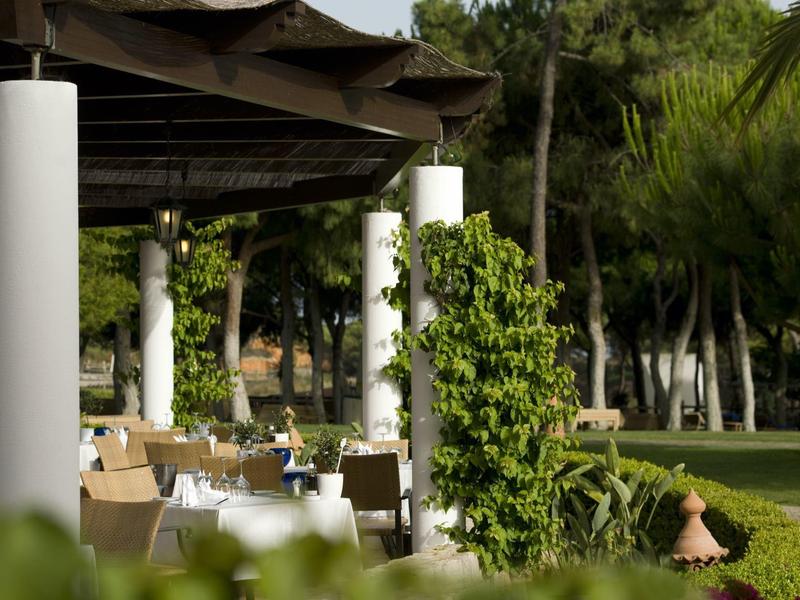 Terrasse mit weißen Säulen, Holzdach, Tischen und Stühlen, umgeben von grünen Pflanzen und Bäumen.